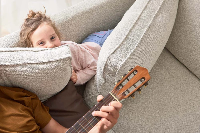 Ein Bild aus dem ADA Shooting auf dem ein Mann auf einem Sofa Gitarre spielt, während ein kleines Kind sich lachend hinter dem Sofakissen versteckt. 