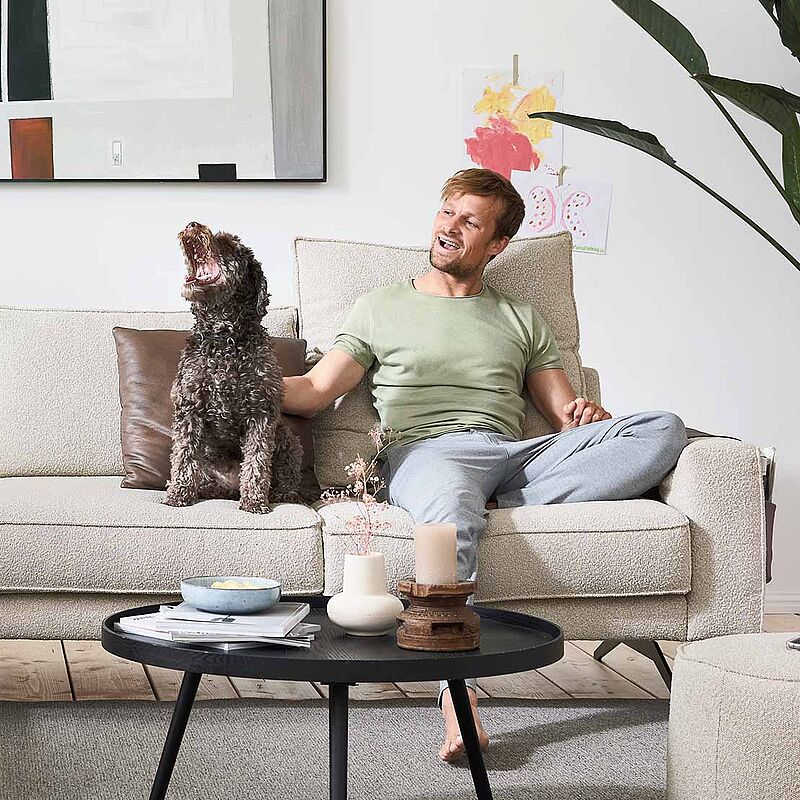 Abgebildet ist ein Wohnzimmer in Nahaufnahme mit einem Mann, der mit seinem Hund auf dem Sofa sitzt. 